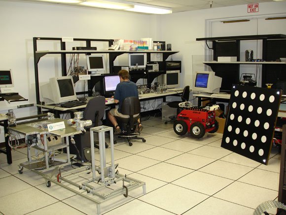 Fig. 2: One view of the workspace in the CLARAty lab showing (left to right) the Dexter rover emulator with manipulator, a software developer at his workstation, two benchtop system including that from Figure 1, the Clifford rover, and a camera calibration target with dots.