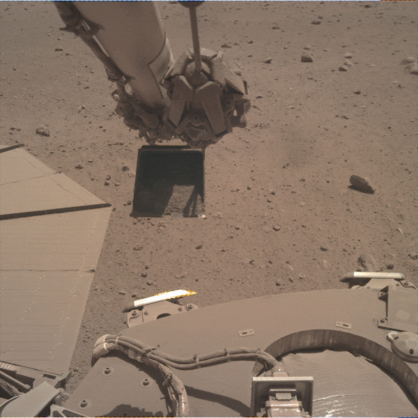 Figure 4: A view from the IDC of the IDA dumping Martian regolith on the lander deck near the West solar array. Dust on the solar array blows away in the wind as sand grains from the dumped regolith impacts the array.